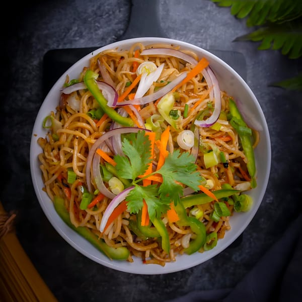 A white bowl of Indian Hakka noodles with carrots, bell peppers, onions, and cilantro on a dark surface.