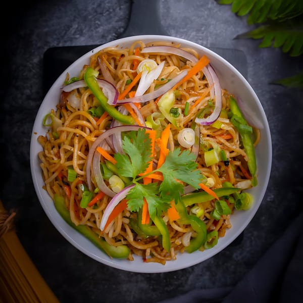 A white bowl of Indian noodles with green peppers, red onions, carrots, and cilantro.