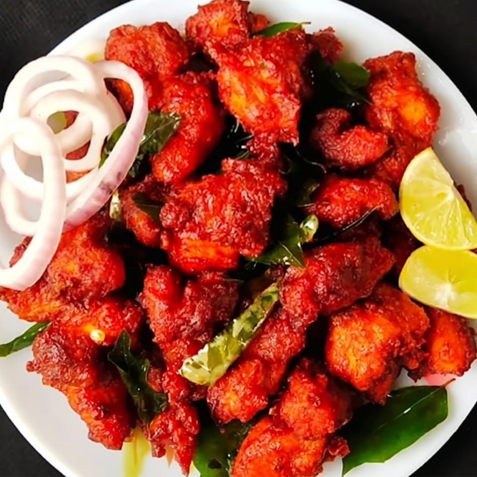 A white plate filled with bright red fried chicken pieces, garnished with onion rings and lime wedges.