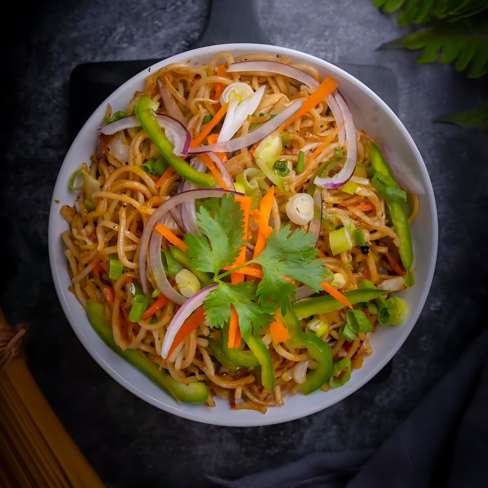 Top-down view of Indian noodles with bell peppers, onions, carrots, and cilantro in a white bowl.