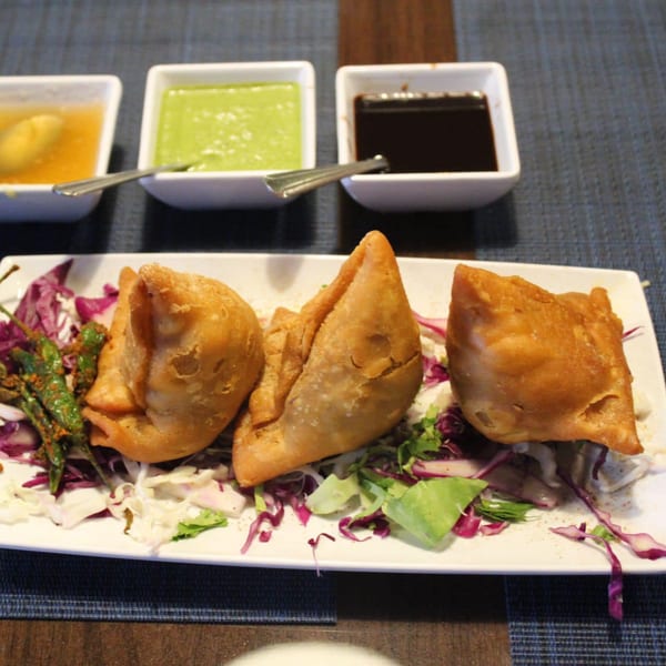 Three golden-brown samosas on a bed of lettuce and red cabbage, with three dipping sauces in the background.